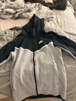 Nike hoodie i svart och grå - Nike hoodie med dragkedja i svart och ljusgrå färg. Hoodien har en stor huva, vita detaljer och Nike-logga på bröstet. Tillverkad i mjukt sweatshirtmaterial, perfekt för chill eller streetwear. Fickor på sidorna och ribbade muddar vid ärmslut och nederkant.