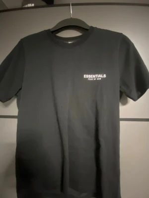 Svart Essentials Fear of God t-shirt - Snygg svart t-shirt från Essentials Fear of God med vit logga på bröstet och stor text på ryggen. Klassisk rund hals och korta ärmar. Perfekt för dig som gillar streetwear och enkel stil.