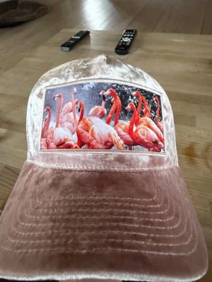 Rosa keps med flamingotryck - Snygg rosa keps i sammetsliknande material med ett stort tryck av flamingos framtill. Kepsen har mesh på baksidan för extra ventilation och justerbar passform. Perfekt för dig som vill sticka ut med en unik accessoar.