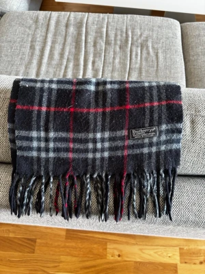 Burberry halsduk - Klassisk halsduk från Burberrys i svart, grått och rött rutmönster. Tillverkad i mjuk ull med tvinnade fransar i ändarna och en snygg vävd logotyp. Perfekt accessoar för att lyfta din outfit runt höst och tidig vår. Har tyvärr inte kvar något från köpet som till exempel låda eller liknande men kontakta gärna mig vid eventuella frågor eller liknande! 