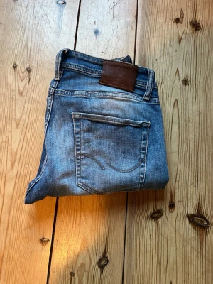 Blå tvättade jeans med skinnpatch - Säljer ett par klassiska blå jeans med snygg tvättad look och raka ben. Jeansen har två bakfickor med dekorativa sömmar och en brun skinnpatch i midjan. Perfekta för en avslappnad stil och passar till det mesta i garderoben. De är några små fläckar på högra bakfickan men de är inget man lägger märke till. Hör av dig vid frågor🙌🏼