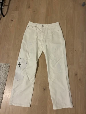 Vita raka jeans med korsdetaljer ChromeHearts - Unika vita jeans med raka ben och dekorativa korsdetaljer i metall på vänster ben. Jeansen har hög midja och coola sydda mönster längs benen som ger en edgy vibe. Perfekta för dig som vill sticka ut med din stil.