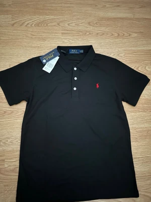 Svart pikétröja från Polo Ralph Lauren - Klassisk svart pikétröja från Polo Ralph Lauren med röd broderad logga på bröstet. Tröjan har korta ärmar, krage och tre knappar framtill. Tillverkad i bomull för en bekväm och stilren look.