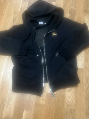 Svart hoodie från Burberry med dragkedja - Svart hoodie från Burberry i storlek M med dragkedja framtill och huva. På bröstet finns en liten guldfärgad Burberry-logga. Hoodien har ribbade muddar och är tillverkad i mjukt material, perfekt för en avslappnad stil.