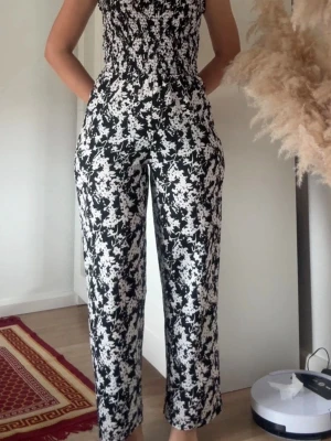 Svartvit jumpsuit från Jacqueline de Yong - Säljer en svartvit ärmlös jumpsuit från Jacqueline de Yong med smockad överdel och raka ben. Mönstret är blommigt och materialet känns lätt och bekvämt, perfekt för varma dagar. Supersnygg och enkel att styla med sneakers eller sandaler.