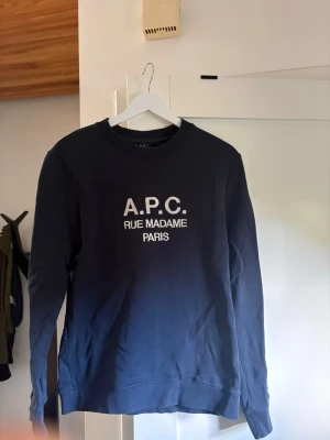 Mörkblå sweatshirt från A.P.C. - Snygg mörkblå sweatshirt från A.P.C. med vit text 'A.P.C. RUE MADAME PARIS' på bröstet. Klassisk rund halsringning och ribbade muddar vid ärmslut och nederkant. Perfekt för en clean och stilren look.