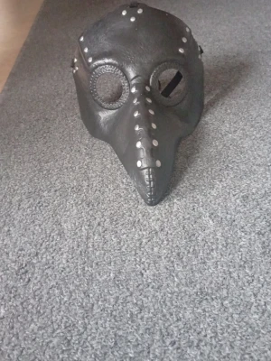 Svart pestdoktorsmask med nitar - Cool svart pestdoktorsmask i konstläder med silverfärgade nitar längs kanterna och näbben. Masken har runda ögonhål med nät och en lång, spetsig näbb. Perfekt för maskerad, cosplay eller halloween.
