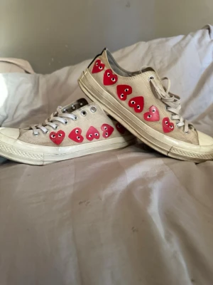 CDG CONVERSE  - Säljer ett par beige sneakers med vita snören och sula. Skorna har flera röda hjärtan med ögon längs sidan som ger en unik och lekfull look. Klassisk låg modell i canvasmaterial som passar perfekt till streetwear och casual outfits.