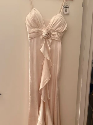 Vit klänning med volangdetaljer - Elegant vit långklänning med smala axelband, v-ringad urringning och dekorativ volang framtill. Klänningen har en markerad midja med en tygblomma och är tillverkad i ett mjukt material som ger en lyxig känsla. Klänningen har aldrig varit använt, endast testad. Nyskick! Storlek S / 36.