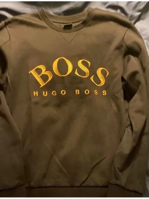 Svart sweatshirt från Hugo Boss - Snygg svart sweatshirt från Hugo Boss med stort gult BOSS-tryck och broderad logga på bröstet. Tröjan har rund halsringning och långa ärmar. Perfekt för dig som vill ha en stilren och trendig look.