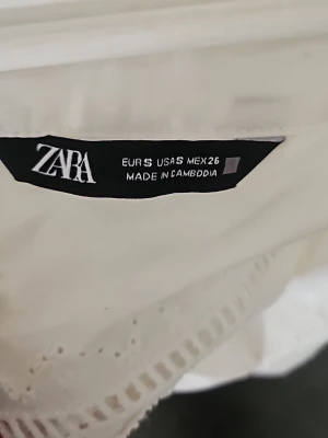 Vit blus med broderade detaljer från Zara - Säljer en vit blus från Zara med fina broderade detaljer och hålmönster längs ärmar och framstycke. Blusen har en luftig känsla och är tillverkad i ett lätt bomullstyg. Perfekt för en fräsch och stilren look.