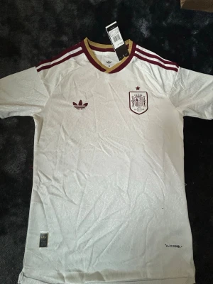 Vit Spanien tröja Adidas - Säljer en vit Spanien tröja från Adidas med vinröda detaljer och guldkrage. Tröjan har Adidas-logga och Spaniens landslagsemblem på bröstet samt diskret mönster i tyget. Perfekt för fotboll och träning.