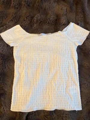 Vit ribbad offshoulder topp - Säljer en ljusvit ribbad topp med offshoulder-design och korta ärmar. Toppen har en stretchig och mjuk struktur som ger en bekväm passform. Strl S men passar även XS. 