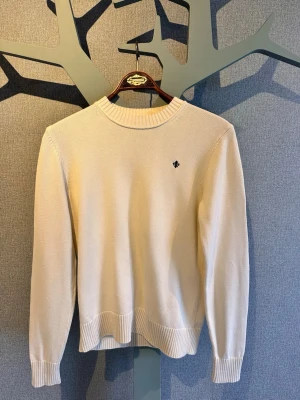 Beige stickad tröja med brodyr - Klassisk beige stickad tröja med rund hals och ribbade muddar vid ärmslut och nederkant. Liten svart broderad detalj på bröstet ger en stilren touch. Perfekt till jeans  för en enkel och snygg look.