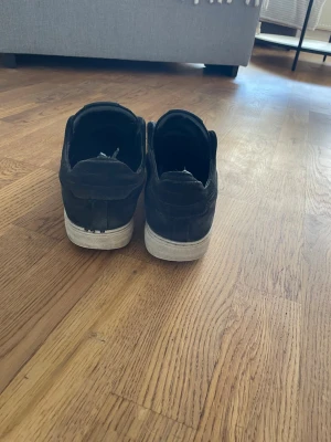 Svarta sneakers från Axel Arigato - Säljer ett par svarta sneakers från Axel Arigato med vit sula och diskret logga på sidan. Skorna har en klassisk låg modell och är tillverkade i mocka med elastiska band över vristen istället för snören. Perfekta för en clean och stilren look. Dessa skor har ja använt rätt mycket men vet inte varför tejp är ibak,