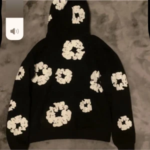 Svart hoodie med vita blommor Denim Tears - Svart hoodie från Denim Tears med stora vita blommor tryckta över hela plagget. Hoodien har en klassisk huva och en avslappnad passform. Perfekt för dig som vill sticka ut med en unik och trendig stil.
