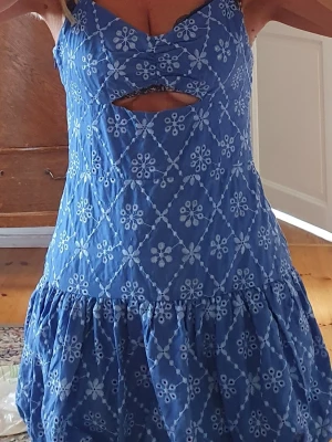 Blå broderad sommarklänning - Söt blå klänning med vita broderade blommor och rutmönster. Klänningen har breda axelband, en trendig cut-out detalj under bysten och volangkjol. Perfekt för varma dagar och har en ungdomlig, fräsch look.