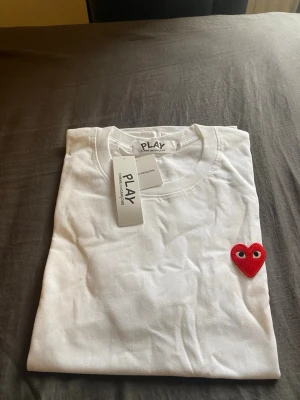 Comme des Garçons T-shirt  - Säljer en vit t-shirt från PLAY med ett ikoniskt rött hjärta med ögon broderat på bröstet. Klassisk rund hals och korta ärmar. Tillverkad i mjuk bomull för en skön känsla. Perfekt för dig som gillar stilrena plagg med en twist.