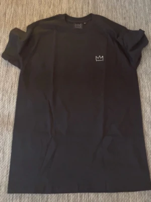 Svart Hov1 t-shirt med turnétryck - Svart t-shirt med liten krona framtill och stort vitt turnétryck på ryggen med texten 'Vi Dansar Vidare I Livet' och turnédatum. T-shirten har rund hals och är i mjukt bomullsmaterial. Perfekt för dig som gillar Hov1 och vill ha en stilren merch.