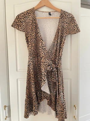 Leopardmönstrad omlottklänning från Divided - Säljer en kort leopardmönstrad omlottklänning från Divided i beige och svart. Klänningen har korta ärmar, knytband i midjan och volangkant nedtill. Perfekt för dig som vill sticka ut med ett trendigt djurmönster.