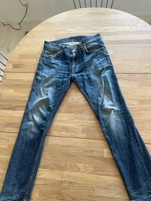 Blå jeans från Nudie Jeans Co - Säljer ett par klassiska blå jeans från Nudie Jeans Co med snygg slitning och kontrastsömmar. Modellen har fem fickor, normal midja och raka ben. Tillverkade i denim med cool tvätt och detaljer i kopparfärg. Perfekta för en avslappnad stil.