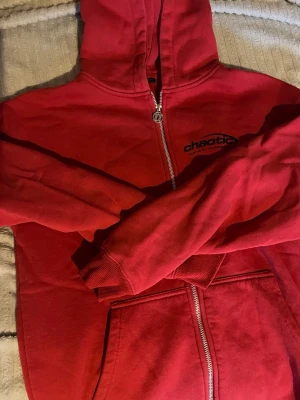 Röd hoodie med tryck från Chaotic - Röd hoodie med dragkedja och huva från Chaotic. Stort svart tryck på ryggen med texten 'chaotic aesthetic awareness from outta space' och mindre logga på bröstet. Mjukt material och fickor framtill. Perfekt för en avslappnad streetstyle.