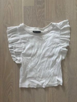 Vit ribbad topp med volangärmar Zara - Supersöt vit ribbad topp från Zara med volangdetaljer på ärmarna. Toppen har rund halsringning och är tillverkad i ett mjukt, stretchigt material som känns skönt mot huden. Perfekt för dig som vill ha en enkel men ändå trendig look. Perfekt till sommaren 😍 