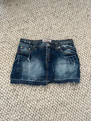 Blå jeanskjol med slitningar - Snygg jeanskjol i kort modell med slitna detaljer och rå kant nertill. Kjolen har klassiska fickor fram och bak samt bälteshällor. Perfekt för en avslappnad och trendig look. I jätte bra skick, använt en gång bara. 37 cm midjemått
