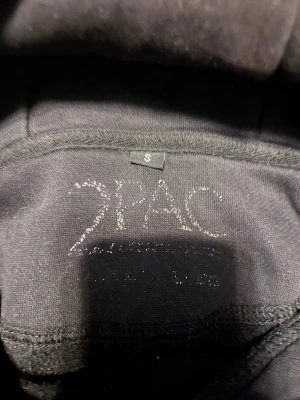 Svart T-shirt med Tupac-tryck - Säljer en svart T-shirt med stort Tupac-tryck på framsidan. T-shirten har klassisk passform och korta ärmar. Materialet känns mjukt och skönt, troligtvis bomull. Perfekt för dig som gillar streetwear och ikoniska motiv. 50kr vid snabbt köp