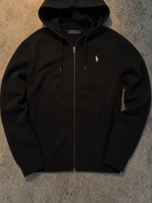 Svart hoodie från Polo Ralph Lauren - Svart hoodie med dragkedja från Polo Ralph Lauren. Klassisk design med vit broderad logga på bröstet, huva med dragsko och två fickor framtill. Tillverkad i mjukt material som passar perfekt för en avslappnad stil.