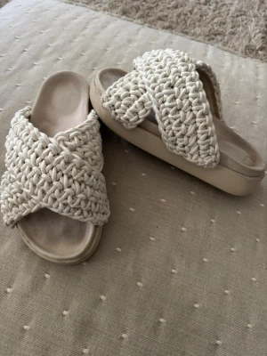 Vita/crem färgade Inuikii flätade slip-in sandaler - Säljer ett par snygga slip-in sandaler med breda, korslagda remmar i beige flätad textil. Sandalerna har en chunky sula och öppen tå, vilket ger en avslappnad och trendig look. Perfekta för sommaren och lätta att matcha med olika outfits.
