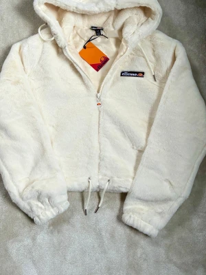 Off white  teddyjacka från Ellesse med huva - Supermjuk och fluffig teddyjacka från Ellesse i krämvit färg. Jackan har huva, dragkedja framtill, dragsko i midjan och broderad logga på bröstet. Perfekt för kalla dagar när du vill vara både varm och snygg.