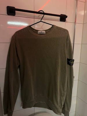 Olivgrön sweatshirt från Stone Island Strl M - Säljer en olivgrön sweatshirt från Stone Island med klassisk rund hals och ribbade muddar. Tröjan har den ikoniska Stone Island-loggan på vänster ärm och är tillverkad i mjukt bomullsmaterial. Perfekt för en avslappnad och stilren look.