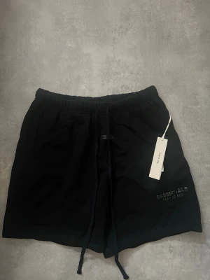 Svarta shorts Essentials Fear of God - Svarta shorts från Essentials Fear of God med elastisk midja och snörning. Klassisk loose fit och diskret logga på vänster ben. Perfekta för chill dagar eller streetwear-stil. Tillverkade i mjuk bomull för maximal komfort.