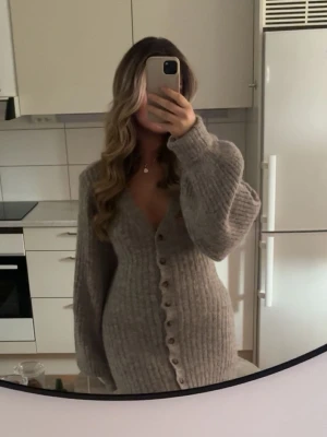 Lång stickad klänning med knappar - Supermysig lång stickad klänning i gråbeige ton med v-ringning och stora ballongärmar. Klänningen har ribbad struktur och knappar hela vägen framtill. Kan även bäras som kofta. Passar S/M/L