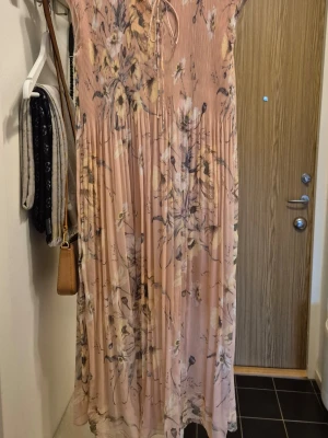 Lång blommig plisserad klänning - Säljer en lång plisserad klänning i ljus aprikos med blommigt mönster i dova toner. Klänningen har v-ringning med snörning framtill och med små knappar och är ärmlös. Kortare foder. Perfekt för sommarens alla tillfällen!