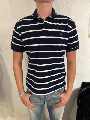 Ralph Lauren Pike - Ralph Lauren pike som är marinblå lite svårt att se på bilderna men den är inte svart. Helt ny och oanvänd, Storleken är custom fit passar mer som L/M. Passar till alla outfits. Skriv om ni har frågor!