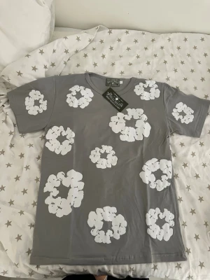 Grå t-shirt med vita blommor - Snygg grå t-shirt med stora vita blommor tryckta över hela tröjan. T-shirten har korta ärmar och en klassisk passform. Perfekt för dig som vill sticka ut med ett coolt mönster och en avslappnad stil.