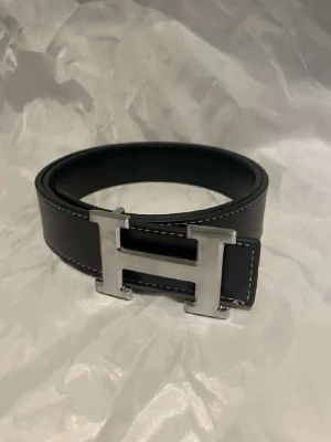 H bälte  - Snyggt svart bälte med ett stort, silvrigt H-format spänne. Bältet har vita sömmar längs kanterna och är gjort i skinn. Perfekt accessoar för att lyfta vilken outfit som helst.