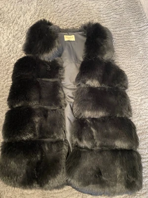 Svart fluffig pälsväst - Snygg svart pälsväst med breda, fluffiga paneler och mjukt foder. Västen har en klassisk krage och är ärmlös, perfekt för att lyfta vilken outfit som helst. Materialet ser ut som fuskpäls och ger en lyxig känsla.