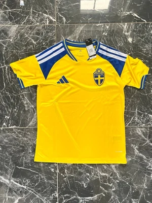 Svensk landslagströja Adidas gul/blå - Gul svensk landslagströja från Adidas med blå detaljer och vita ränder på axlarna. Broderad svensk landslagssköld på bröstet och Adidas-logga i blått. Tillverkad i lätt och ventilerande material, perfekt för träning eller match.