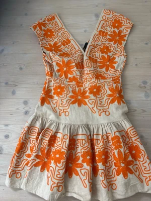 Kort beige klänning med orange broderi - Unik kort klänning i beige linne från Maje med stora orange blommiga broderier. Klänningen har v-ringning, är ärmlös och har en volangkjol.