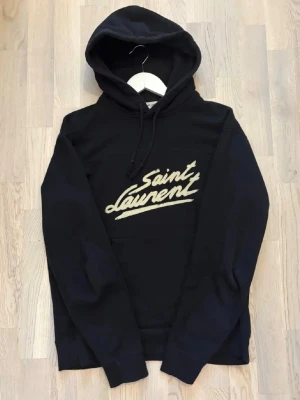 Svart Saint Laurent hoodie - Svart hoodie från Saint Laurent med stor huva och dragsko. Framsidan har ett broderat Saint Laurent-tryck i vitt. Klassisk passform och mjukt material som känns lyxigt. Perfekt för dig som gillar streetstyle och designerplagg.