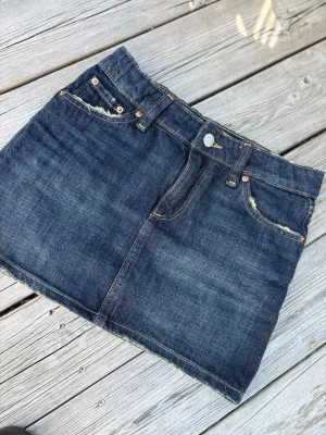 Mörkblå jeanskjol med rå kant - Snygg mörkblå jeanskjol i klassisk femficksmodell med rå, fransig kant nedtill. Kjolen har bälteshällor, knapp och dragkedja framtill samt slitna detaljer vid fickorna för en cool look. Passar mig som är 160 cm. En jeanskjol i taight modell, lite ”längre modell” om man jämför hur korta många av dagens kjolar är! 🤗