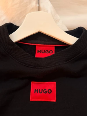 Svart sweatshirt från HUGO - Svart sweatshirt från HUGO med röd logotyp-patch på bröstet. Tröjan har rund halsringning, långa ärmar och ribbade muddar vid ärmslut och nederkant. Perfekt för en stilren och modern look.