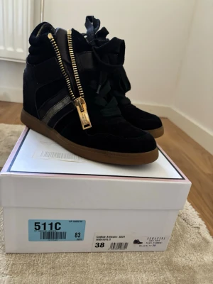 Serafini Manhattan skor - Snygga svarta high top sneakers från Serafini med guldfärgade dragkedjor och spännen. Skorna har svarta mockadetaljer, svarta snören och en brun gummisula. Perfekta för dig som vill ha en unik och trendig look.