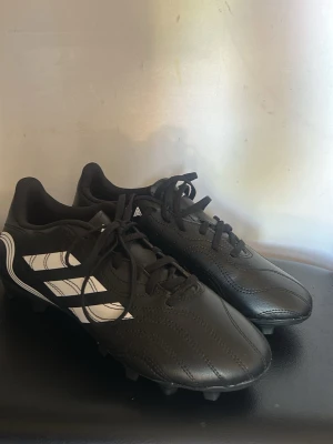 Svarta Adidas fotbollsskor med dobbar - Säljer ett par klassiska svarta Adidas fotbollsskor med vita ränder på sidan. Skorna har snörning och slitstark yttersula med fasta dobbar för bra grepp på planen. Tillverkade i syntetmaterial och designade för att ge stabilitet och komfort under match och träning.