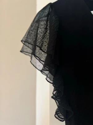 Svart topp med volangärmar - Snygg svart topp med v-ringning och stora, transparenta volangärmar i mesh. Perfekt för dig som vill ha något extra till outfiten. Toppen har en figurnära passform och är riktigt trendig.