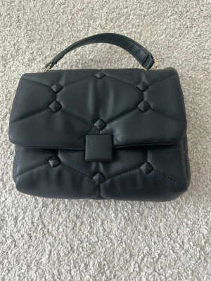Svart  handväska Zara - Snygg svart handväska i quiltad design med geometriskt mönster. Väskan har ett kort handtag upptill och stängs med ett fyrkantigt spänne. Tillverkad i mjukt konstläder som ger en lyxig känsla. 