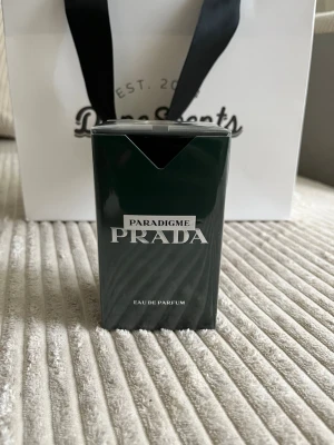 Prada Paradigme Eau de Parfum - Prada Paradigme Eau de Parfum i en elegant mörkgrön rektangulär flaska med silverdetaljer. Förpackningen har en lyxig känsla och är tillverkad av kartong. Perfekt för dig som gillar exklusiva dofter och snygg design.